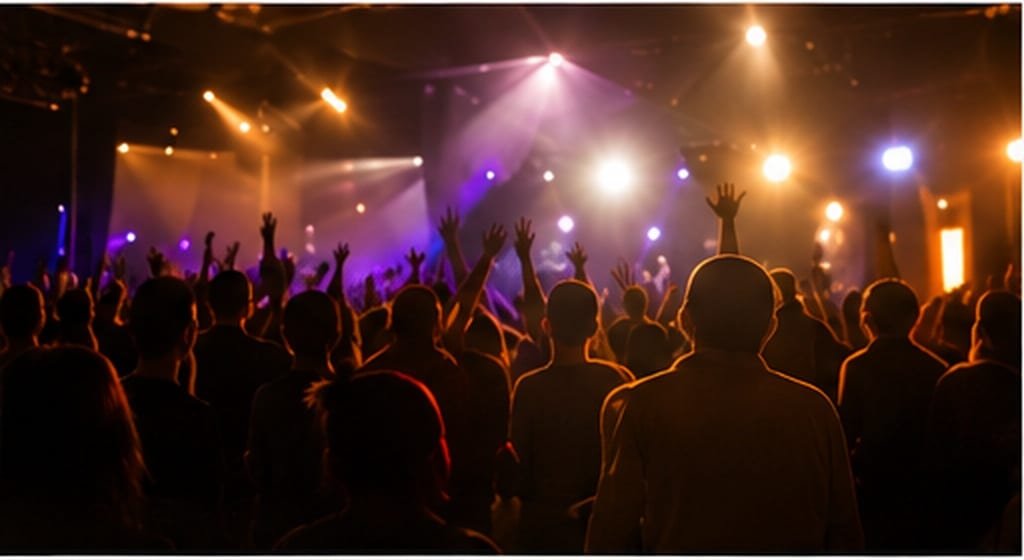 A crowd photographed from behind with many hands raised, lit by purple and amber stage lighting